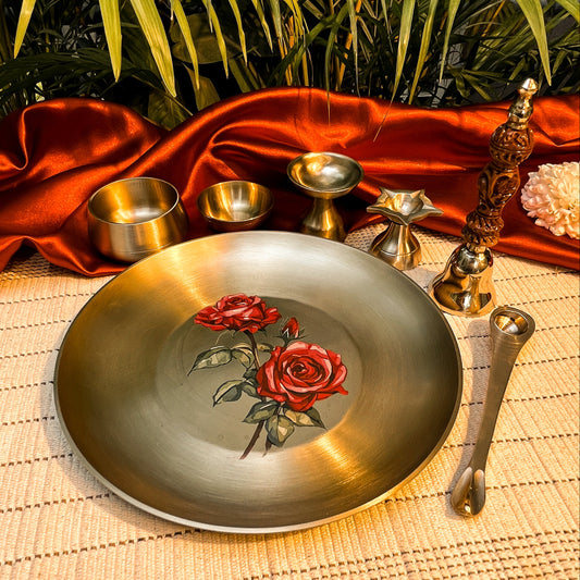 Decorative brass plate with red roses on a textured surface with a red cloth and greenery in the background.